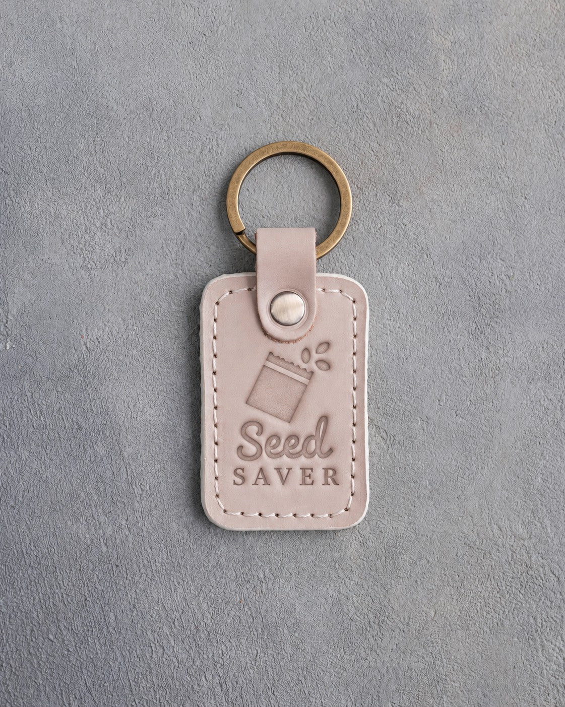 Gray sand leather keychain debossed with an open seed packet above Seed SAVER, on gray concrete