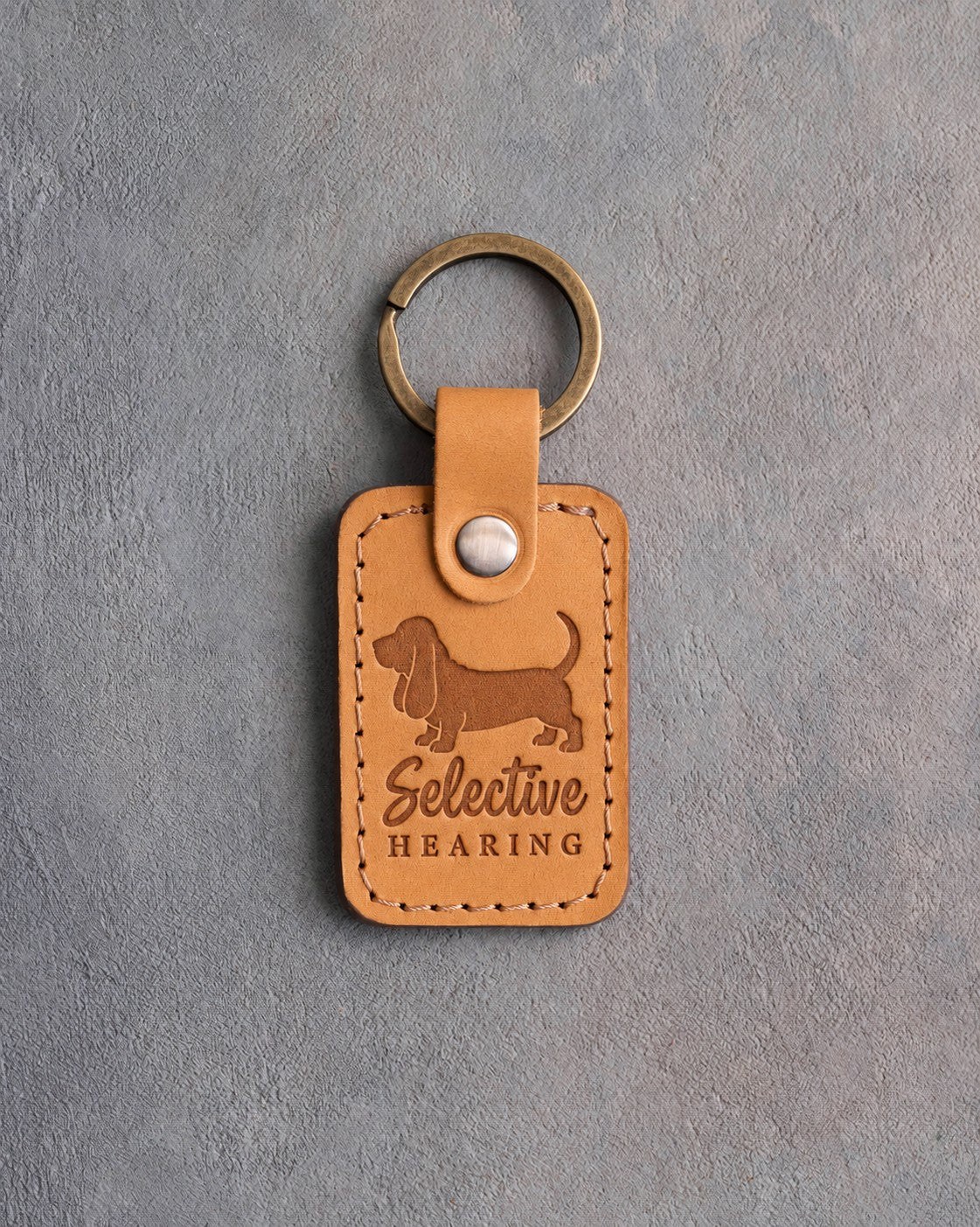 Indian Summer tan leather keychain debossed with a Basset Hound silhouette and the words Selective HEARING, on gray concrete