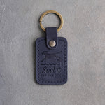 Blueberry navy leather keychain debossed with a dog leaping off a dock and the words Send it OFF THE DOCK, on gray concrete