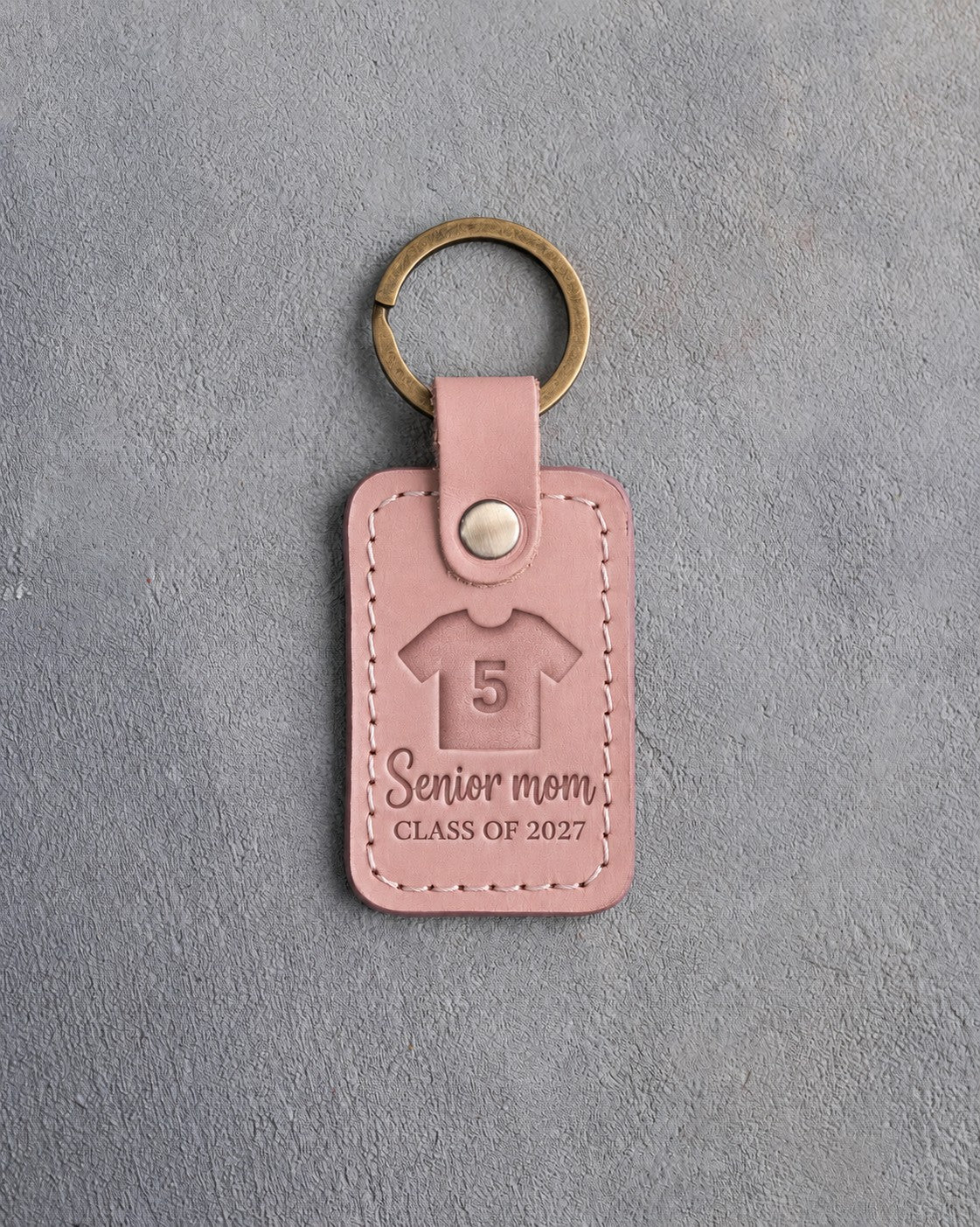 Blush pink leather keychain debossed with a jersey numbered 5 and the words Senior mom CLASS OF 2027, on gray plaster