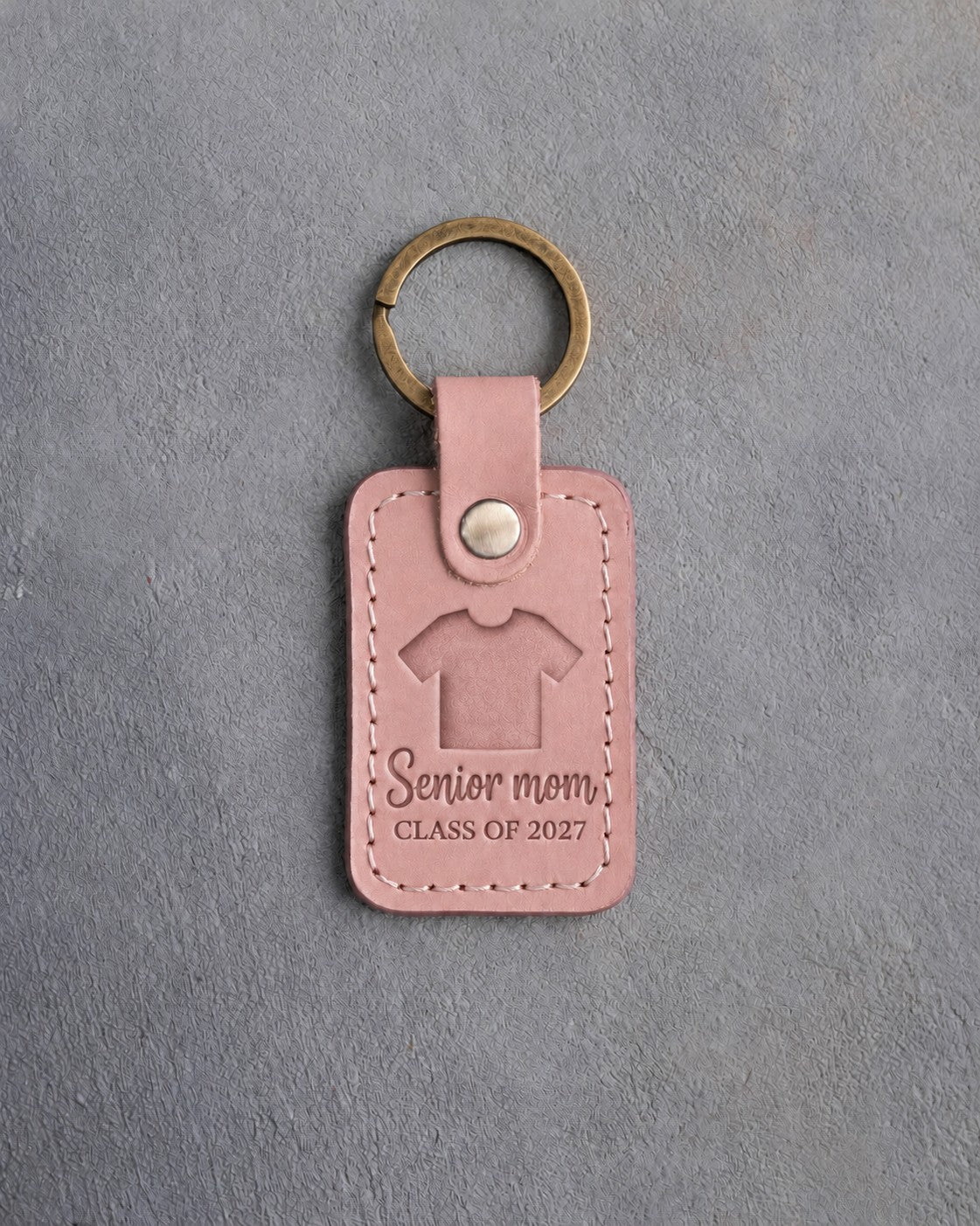 Blush pink leather keychain debossed with a sports jersey above the words Senior mom and CLASS OF 2027, on gray concrete