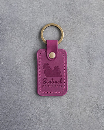 Fuchsia pink leather keychain debossed with a Lhasa Apso silhouette and the words Sentinel OF THE SOFA, on gray concrete