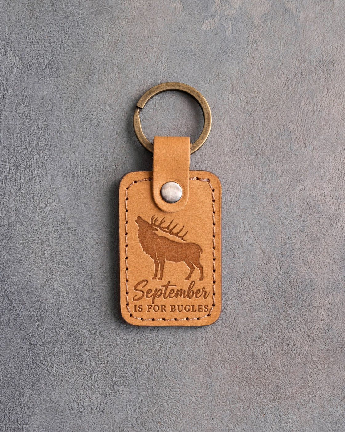 Golden tan leather keychain debossed with a bugling bull elk above the words September IS FOR BUGLES, on gray plaster