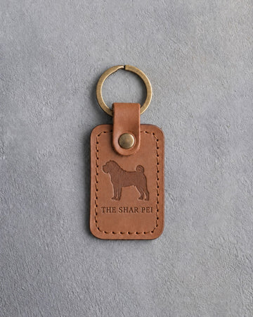 Caramel leather keychain debossed with a Shar Pei silhouette above THE SHAR PEI, on gray concrete