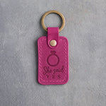 Fuchsia leather keychain debossed with a solitaire engagement ring above the words She said YES, on gray concrete