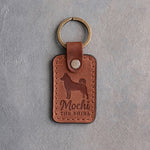 Arizona tan leather keychain debossed with a Shiba Inu silhouette, the name Mochi and THE SHIBA, on gray concrete