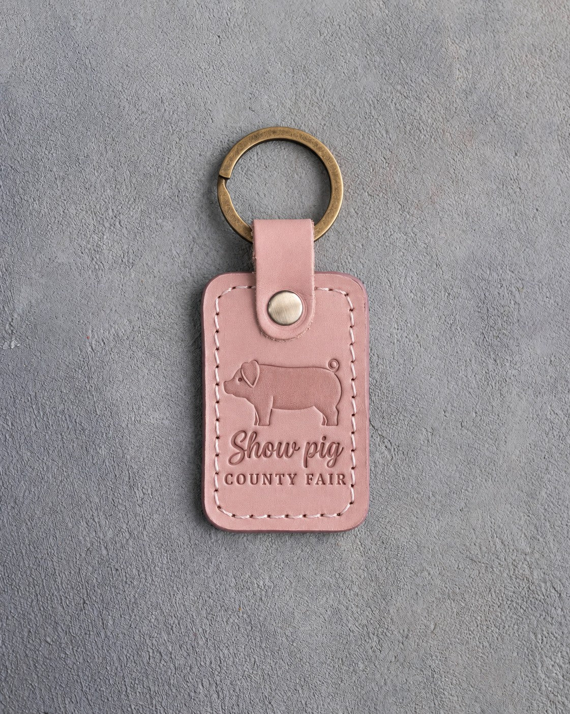 Blush pink leather keychain debossed with a curly-tailed pig above the words Show pig COUNTY FAIR, on gray concrete