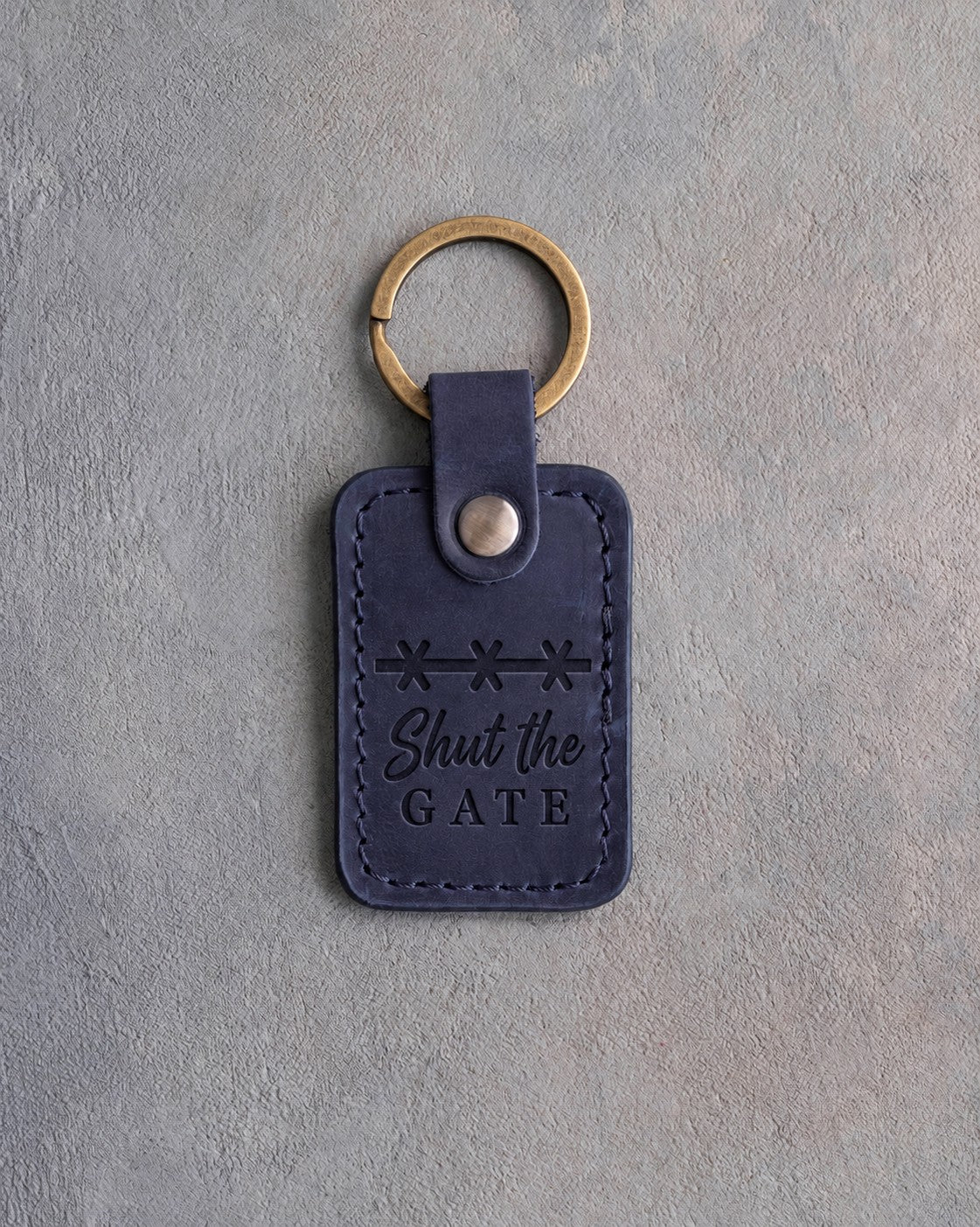Blueberry navy leather keychain debossed with a band of barbed wire above Shut the and GATE, on gray plaster