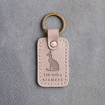 Gray sand leather keychain debossed with a sitting Siamese cat above Life with a and SIAMESE, on gray concrete