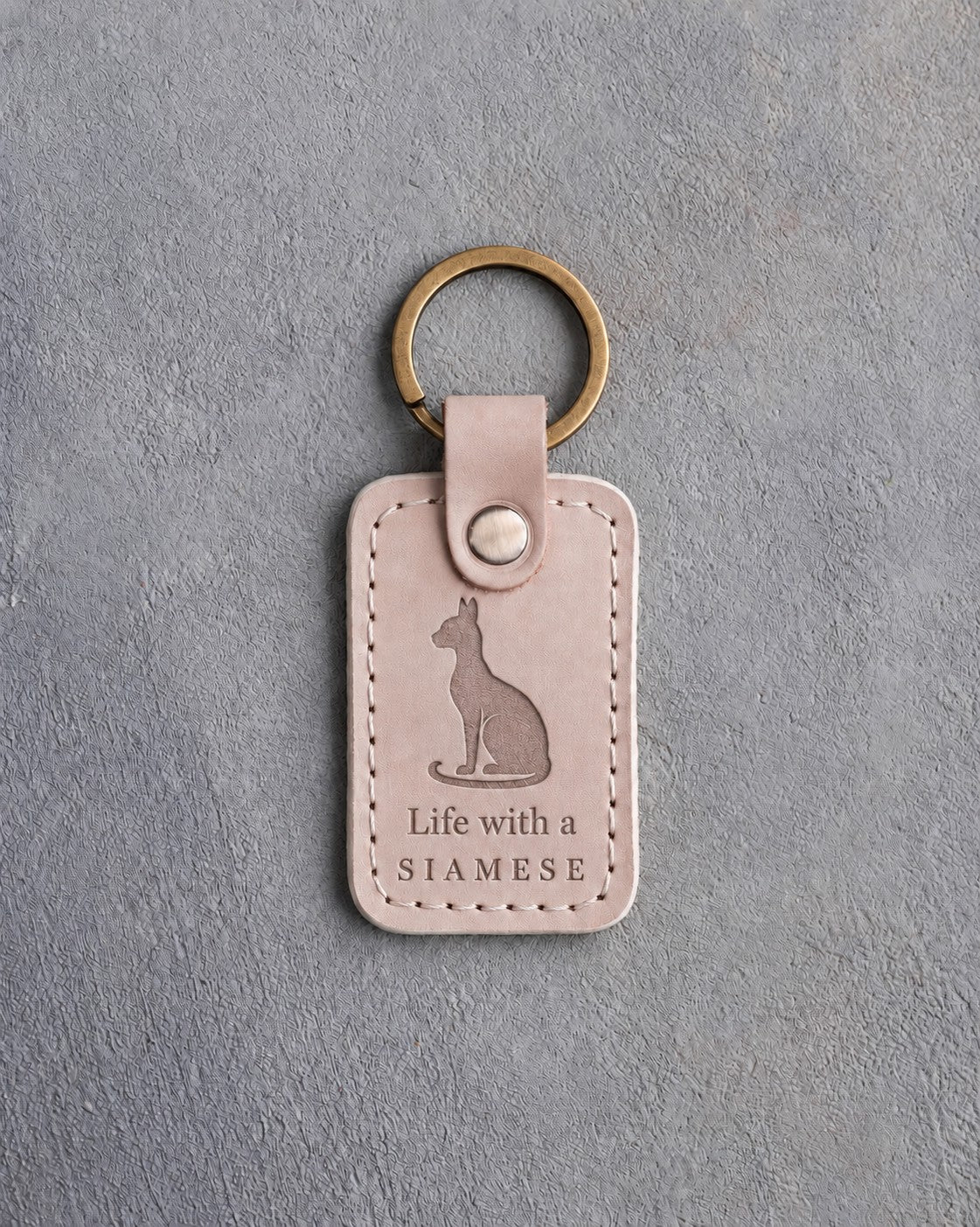Gray sand leather keychain debossed with a sitting Siamese cat above Life with a and SIAMESE, on gray concrete