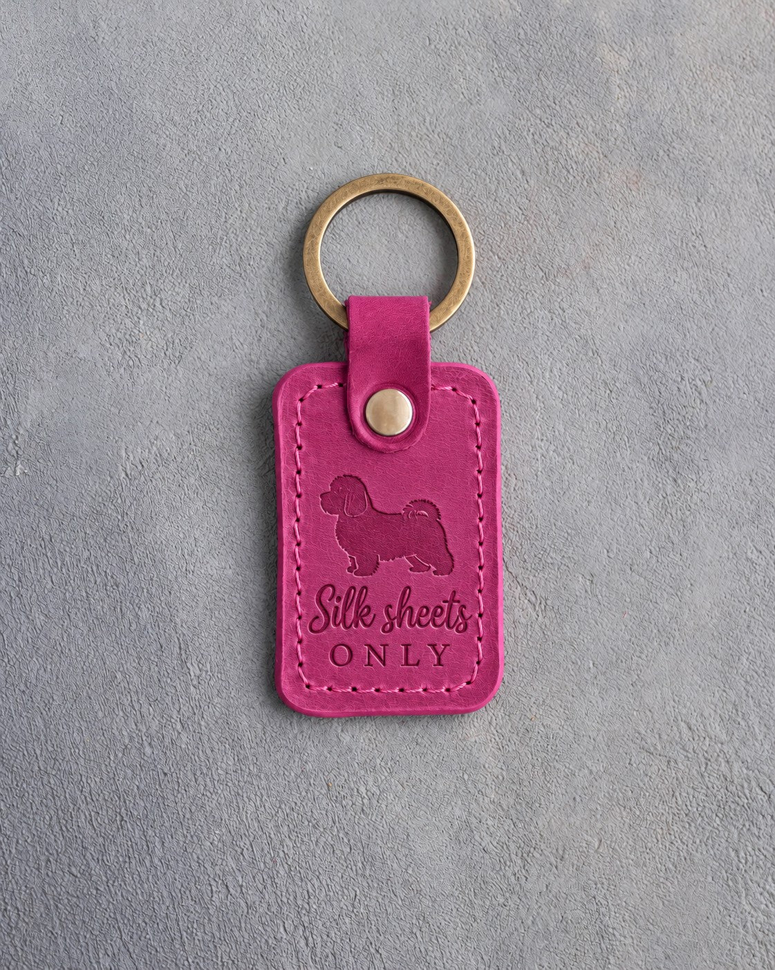 Fuchsia pink leather keychain debossed with a Maltese silhouette and the words Silk sheets ONLY, on gray concrete