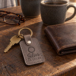 Silver 25 YEARS keychain in gray leather on an oak kitchen table beside two stoneware mugs and reading glasses