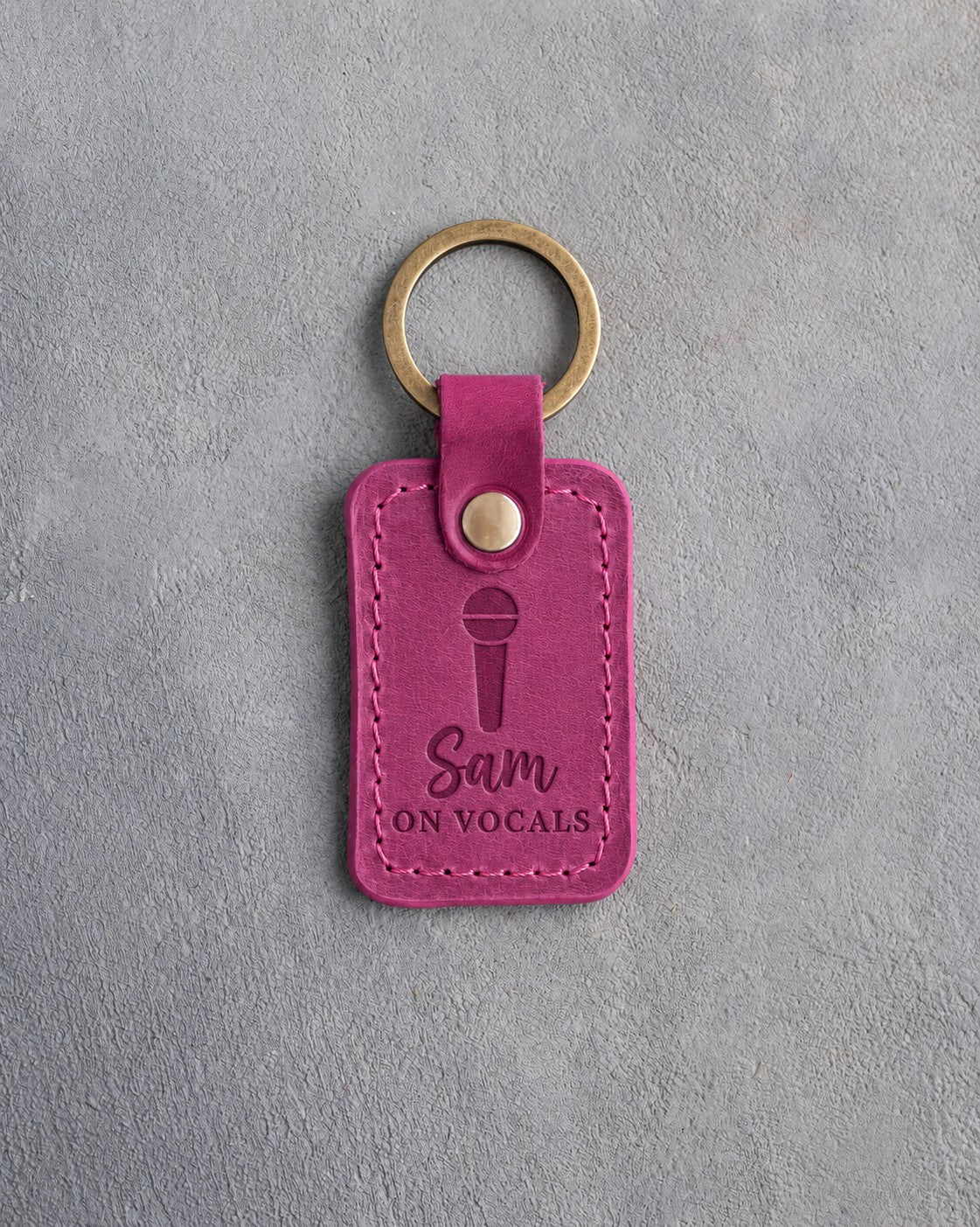 Fuchsia pink leather keychain debossed with a handheld microphone above the name Sam and ON VOCALS, on gray plaster