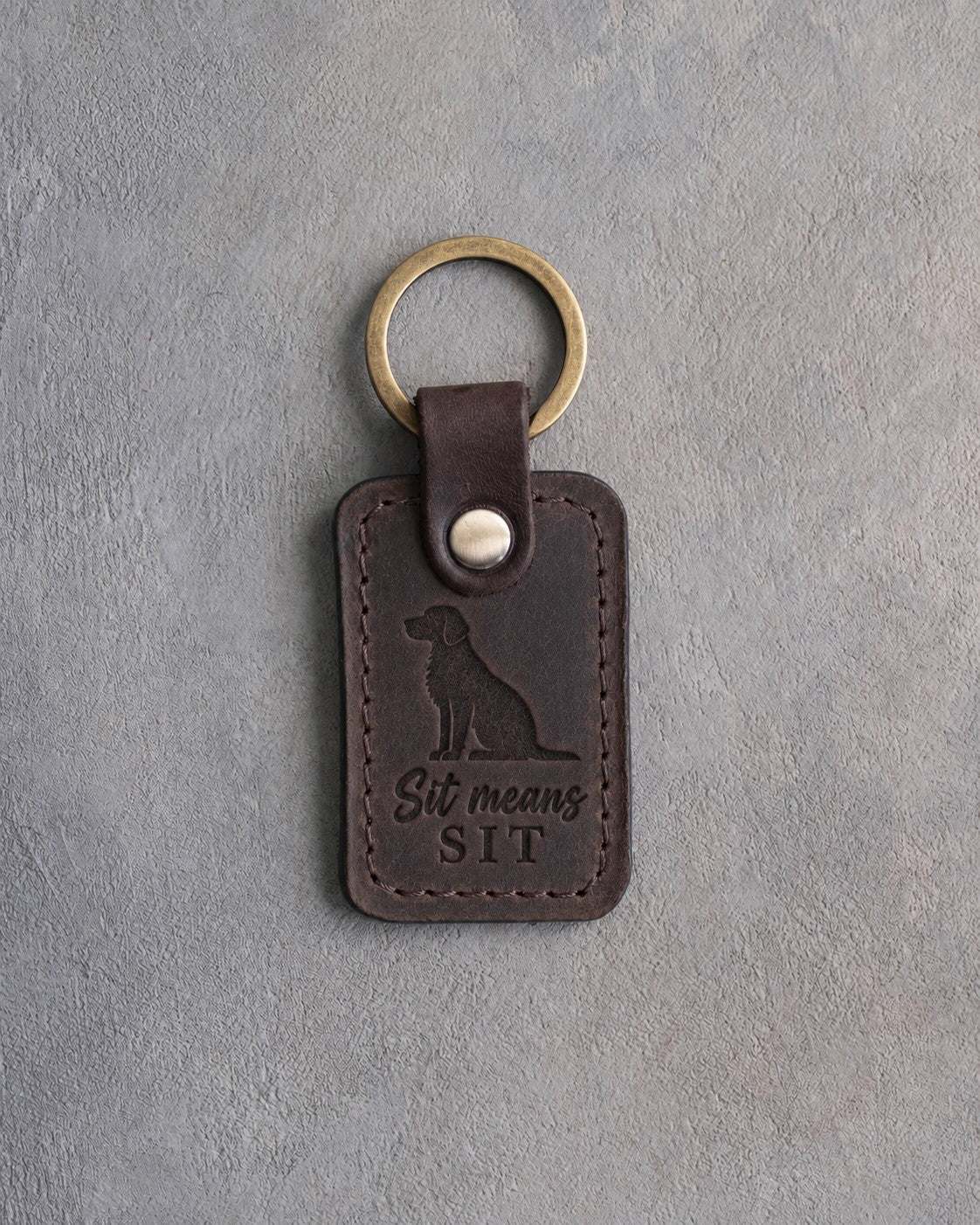 Dark espresso leather keychain debossed with a sitting mixed-breed dog and the words Sit means SIT, on gray concrete