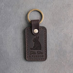 Dark espresso leather keychain debossed with a French bulldog sitting upright on its rump and the words Sits like A PERSON, on gray concrete