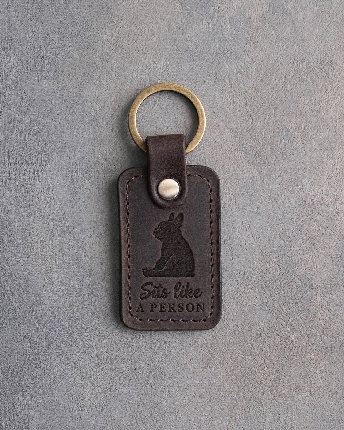 Dark espresso leather keychain debossed with a French bulldog sitting upright on its rump and the words Sits like A PERSON, on gray concrete