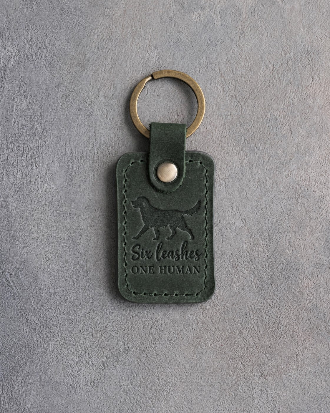 Forest green leather keychain debossed with a trotting dog and the words Six leashes ONE HUMAN, on gray concrete