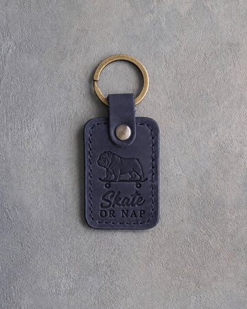 Blueberry navy leather keychain debossed with an English bulldog riding a skateboard and the words Skate OR NAP, on gray concrete