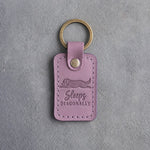 Lilac leather keychain debossed with a dog stretched flat and the words Sleeps DIAGONALLY, on gray concrete