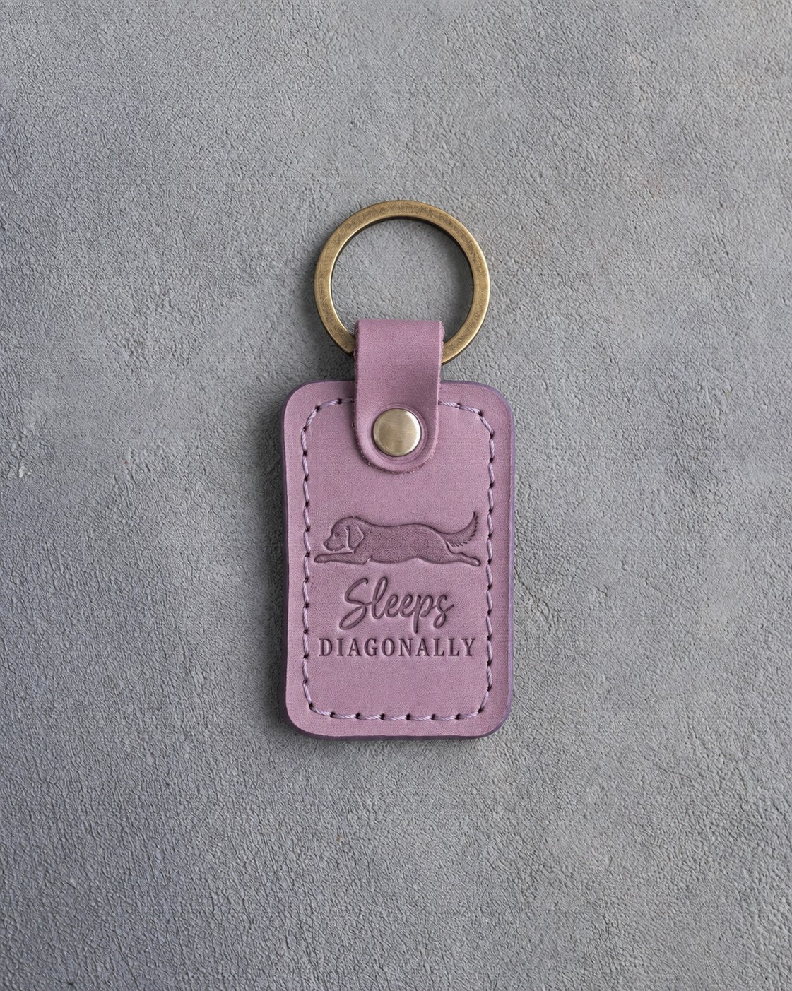 Lilac leather keychain debossed with a dog stretched flat and the words Sleeps DIAGONALLY, on gray concrete