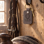 Country gray Smells like HORSE keychain on a forged bridle hook against rough pine boards in a tack room