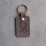 Country gray leather keychain debossed with a horseshoe above Smells like and HORSE, on gray plaster