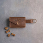 Brown leather dog treat pouch with a snap flap stamped BUDDY, a strap with a D clip and a few small treats beside it