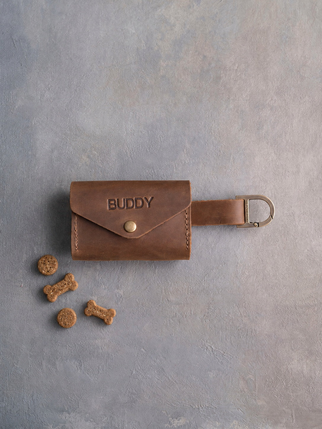 Brown leather dog treat pouch with a snap flap stamped BUDDY, a strap with a D clip and a few small treats beside it