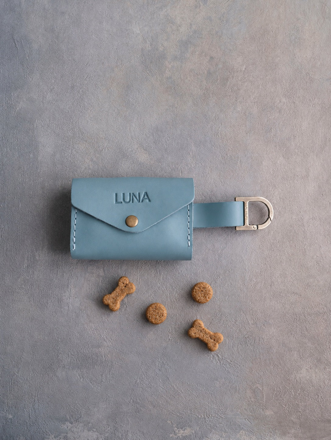 Light blue leather dog treat pouch with a snap flap stamped LUNA and small dog treats on grey plaster