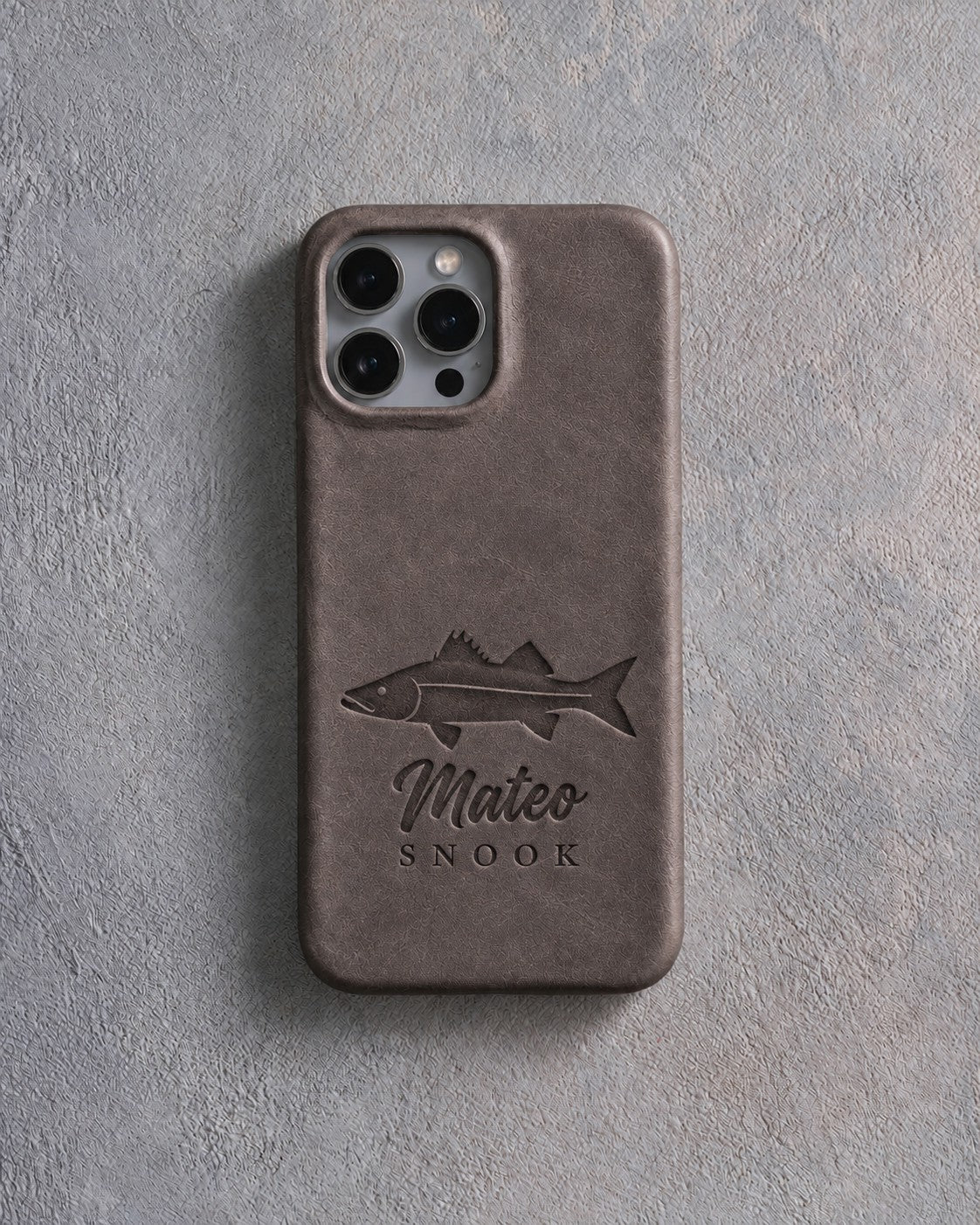 Back of a country gray leather iPhone case debossed with a snook above Mateo and SNOOK below the camera, on gray plaster