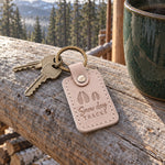 Snow Day Tracks keychain in pale gray leather resting on fresh snow beside a child's red mitten
