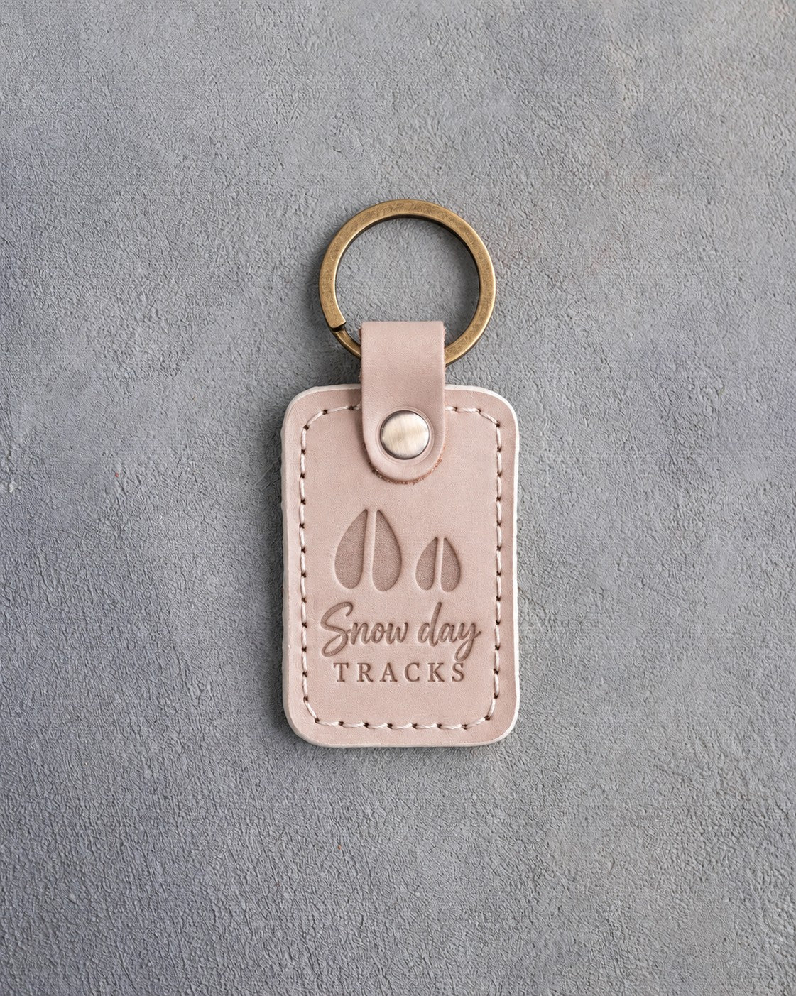 Gray sand leather keychain debossed with a large deer track and a small fawn track above the words Snow day TRACKS, on gray plaster