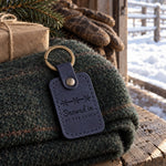Blueberry Snowed in AT THE RANCH keychain on a folded green wool blanket inside a half-open barn door, snow deep on the corral rails outside