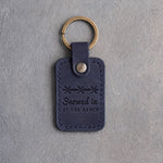 Blueberry navy leather keychain debossed with a band of barbed wire above Snowed in and AT THE RANCH, on gray plaster