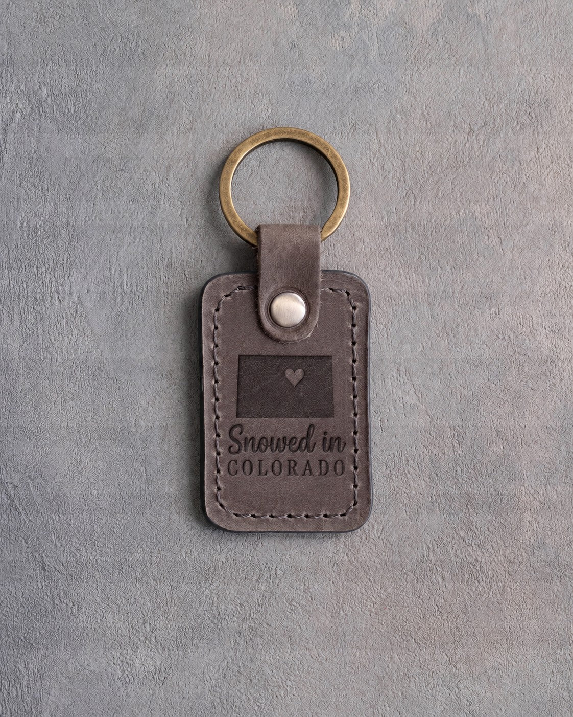 Country gray leather keychain debossed with a solid Colorado rectangle and a heart at Denver above Snowed in and COLORADO, on gray concrete