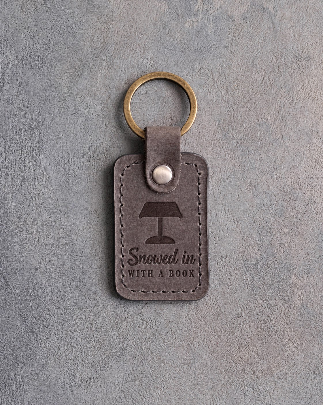 Country gray leather keychain debossed with a banker's lamp above Snowed in and WITH A BOOK, on gray concrete