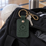 Green Soccer Dad keychain clipped to a mesh ball bag on a grass field on a cool morning
