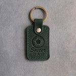 Forest green leather keychain debossed with a soccer ball and the words Soccer DAD, on gray plaster