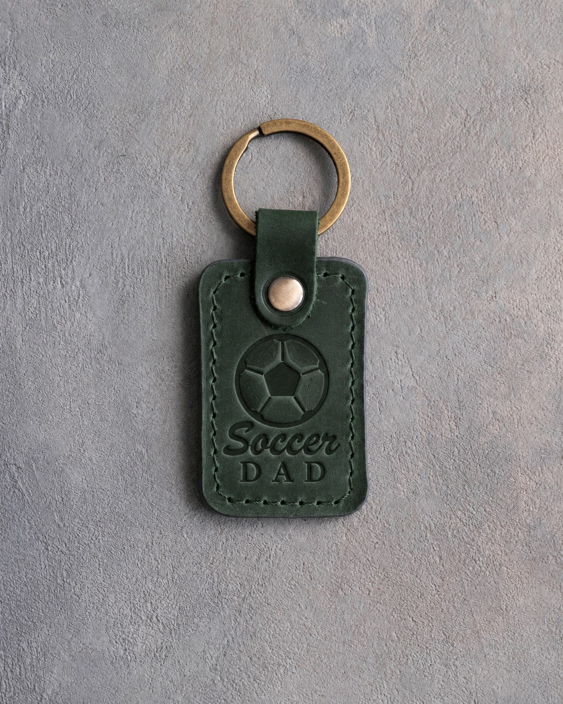 Forest green leather keychain debossed with a soccer ball and the words Soccer DAD, on gray plaster