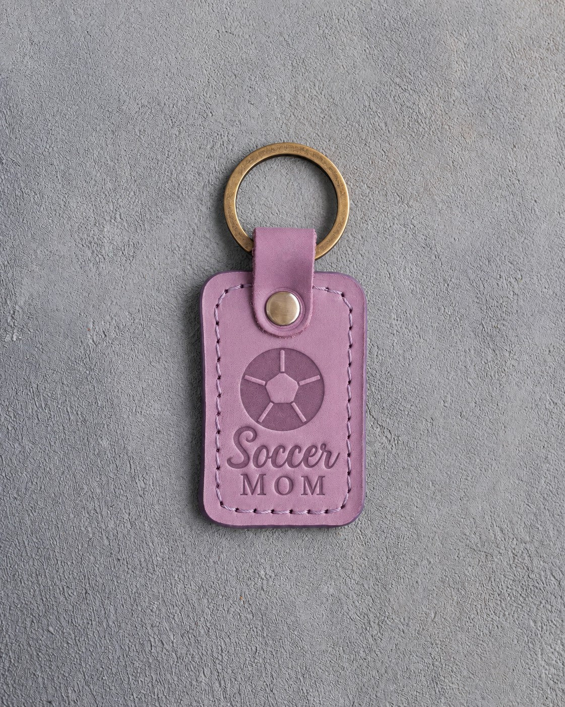 Lilac leather keychain debossed with a soccer ball and the words Soccer MOM, on gray plaster