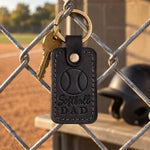 Black Softball Dad keychain hanging on a chain-link dugout fence in low afternoon sun