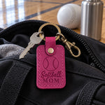 Bright pink Softball Mom keychain clipped to a bat backpack hanging on a dugout fence