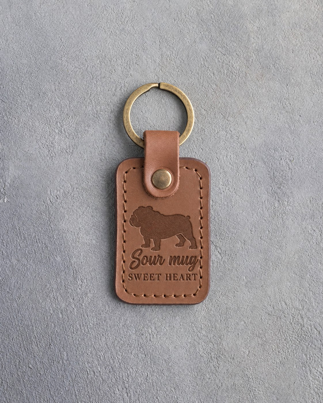Caramel leather keychain debossed with an English bulldog silhouette and the words Sour mug SWEET HEART, on gray concrete
