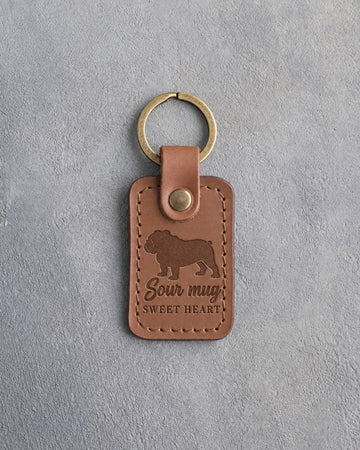Caramel leather keychain debossed with an English bulldog silhouette and the words Sour mug SWEET HEART, on gray concrete