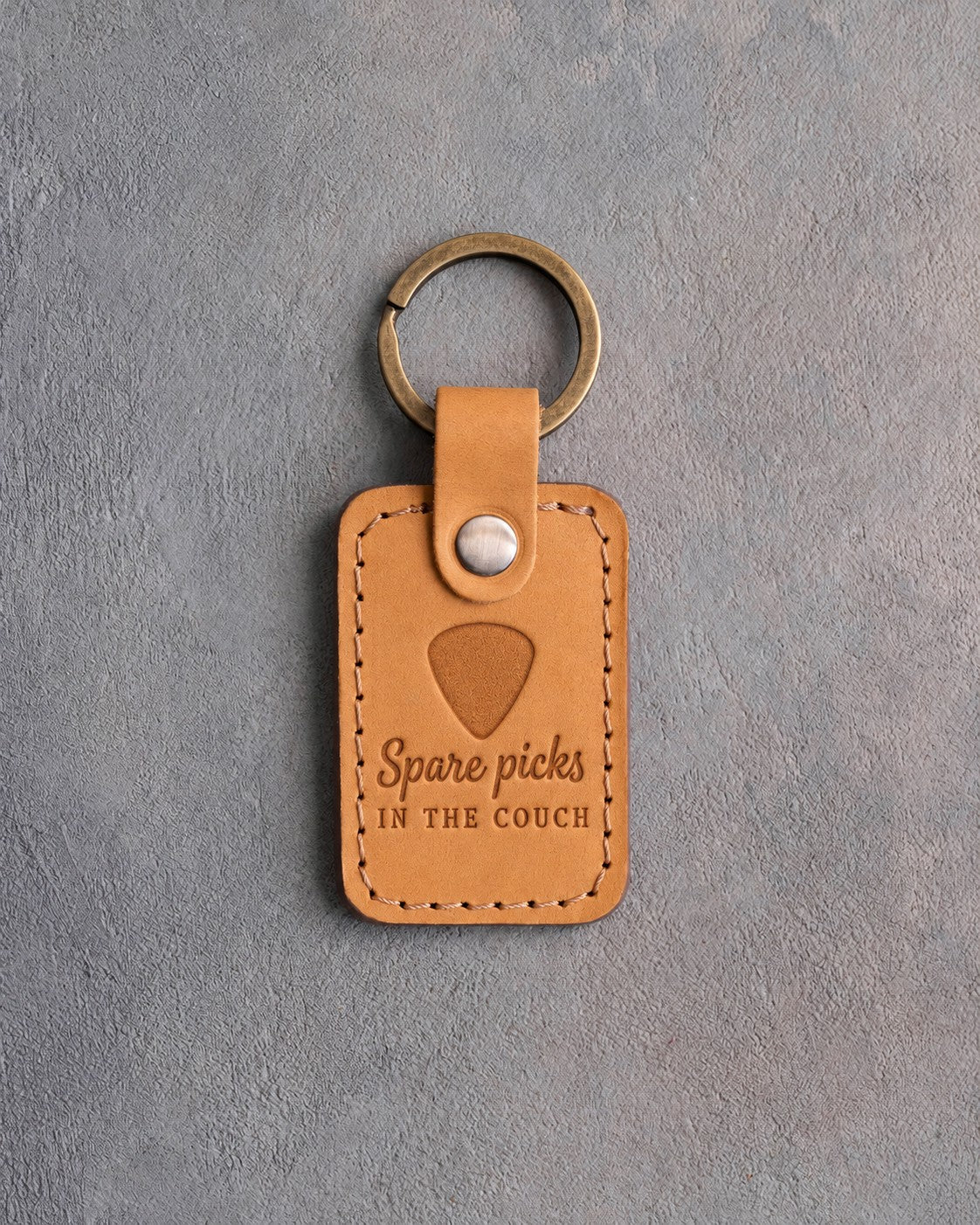 Golden tan leather keychain debossed with a guitar pick above the words Spare picks IN THE COUCH, on gray plaster