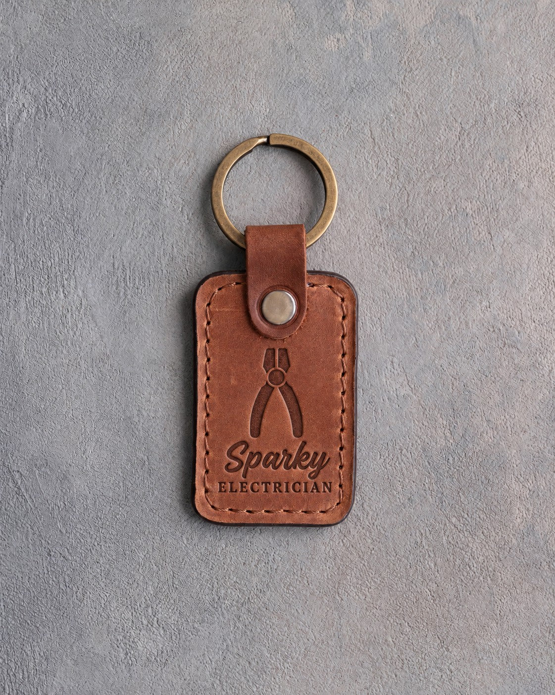 Copper-toned leather keychain debossed with upright lineman's pliers above the words Sparky and ELECTRICIAN, on gray plaster