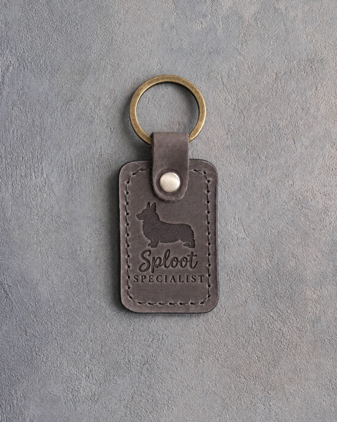 Country gray leather keychain with a debossed corgi silhouette and the words Sploot SPECIALIST on gray concrete