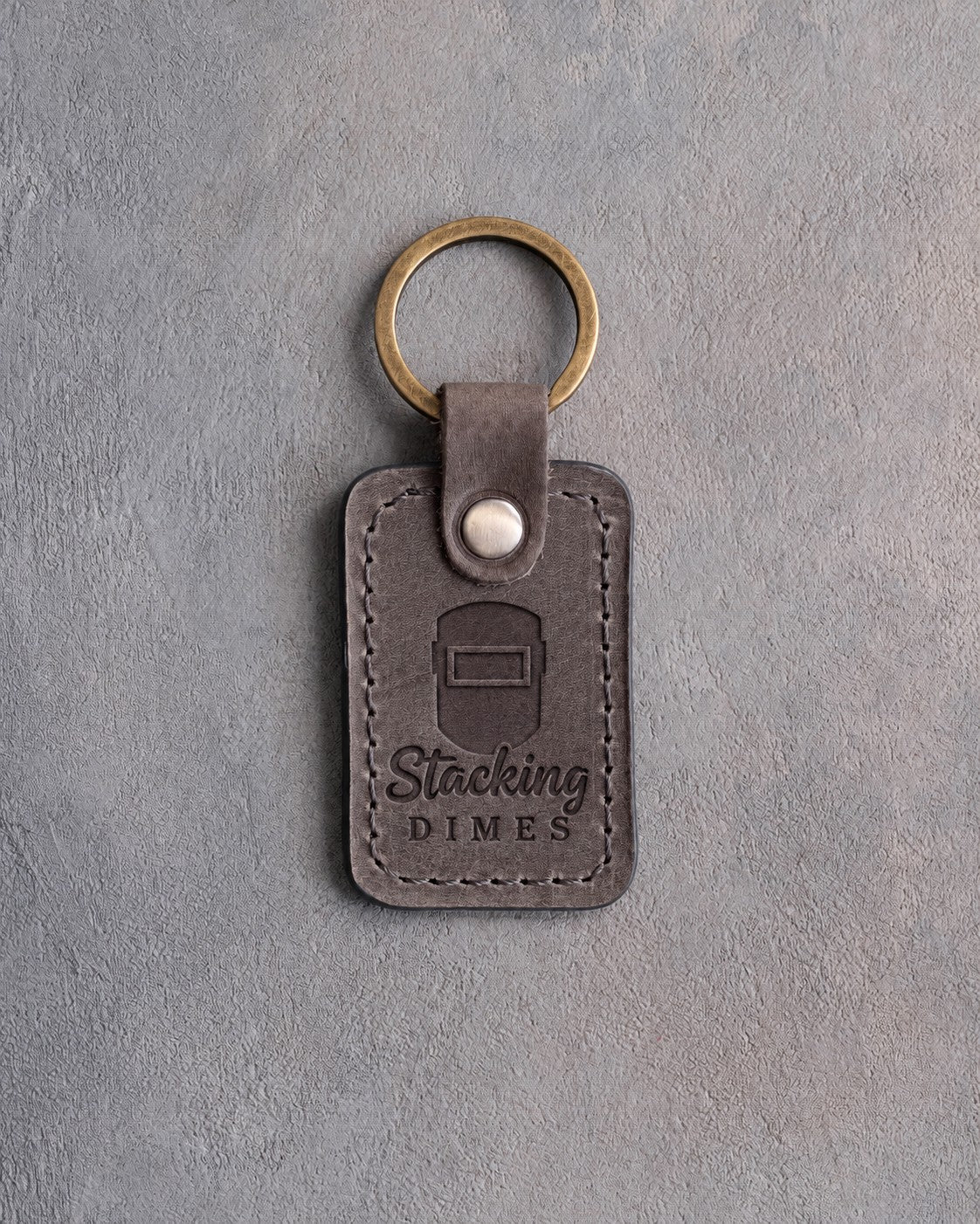 Country gray leather keychain debossed with a welding hood above the words Stacking and DIMES, on gray plaster
