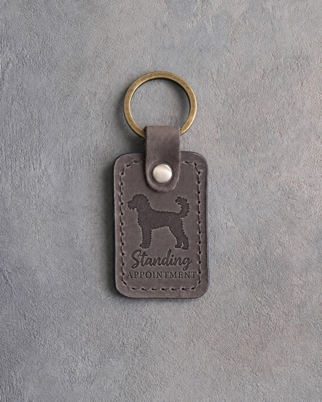Country gray leather keychain debossed with a curly goldendoodle silhouette and the words Standing APPOINTMENT, on gray concrete