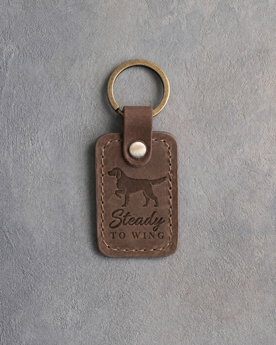 Sicilian brown leather keychain debossed with a pointing dog and the words Steady TO WING, on gray concrete